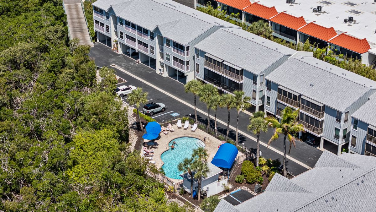 Aerial view of the resort property showing multi-story buildings with balconies, a central swimming pool with blue umbrellas, and lush tropical landscaping.